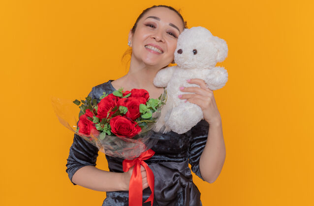 国际快乐的亚洲女人拿着一束红玫瑰和泰迪熊作为礼物 站在橘色的墙上欢笑着庆祝母亲节亚洲快乐微笑