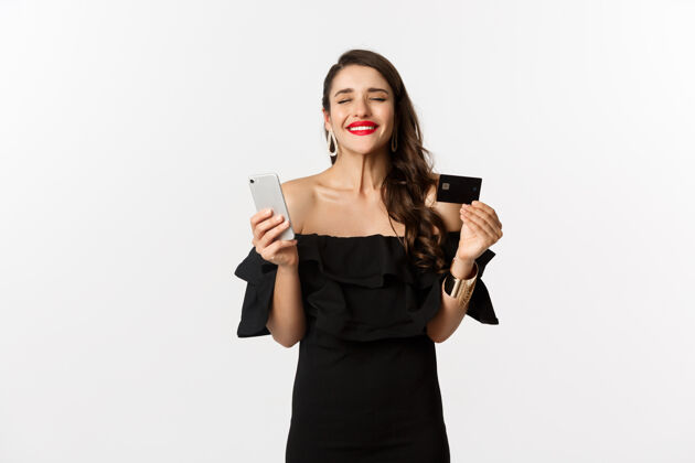 服饰网上购物概念穿着黑色衣服的时髦女人 拿着智能手机的信用卡 看起来很满意 站在白色背景下魅力年轻女人