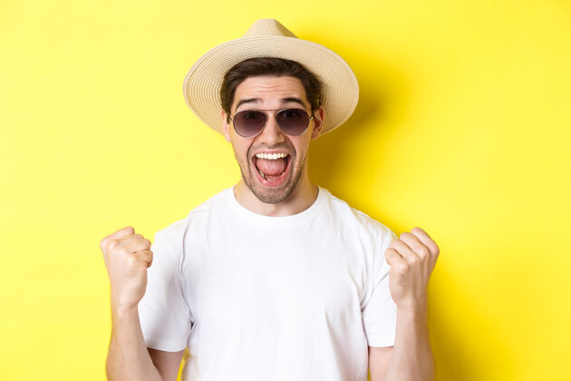 黄色旅游和度假的概念幸运的人旅游者赢得门票 做拳头泵和说是的 穿着度假服 站在黄色背景下夏天男人帽子