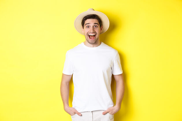 年轻惊讶的男子戴着草帽的游客看起来很高兴 对旅行社的广告反应惊讶 站在黄色的背景上成人男人男人