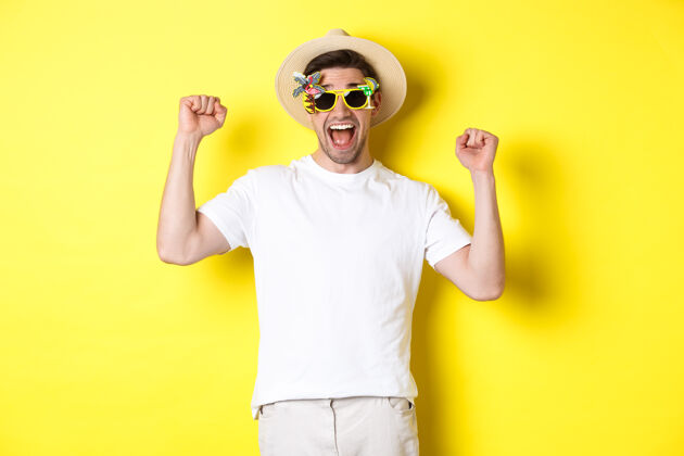 帅气旅游和生活方式的概念快乐的人赢得旅游胜地之旅 喊是的 举手 胜利 戴墨镜和夏季帽子 黄色背景帽子夏天自信