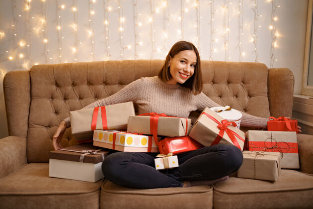 黑发幸福的年轻女子抱着许多礼物盒盘腿坐在一个驼色沙发与灯拥抱梦想闪亮