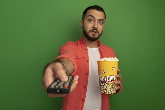 电视身穿橙色衬衫 留着胡子的年轻人拿着装有爆米花的水桶 用电视遥控器站在绿色的墙上 看上去很惊讶年轻人使用桶