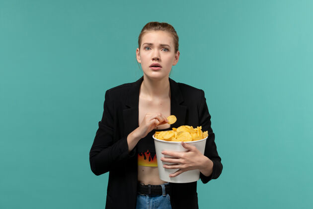 看正面图：年轻女性在淡蓝色的表面上吃薯片看电影抱土豆前面