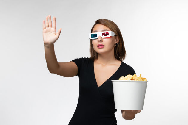 前面正面图：戴着d型太阳镜手持薯片的年轻女性 在白色表面挥手致意漂亮女商人电影院