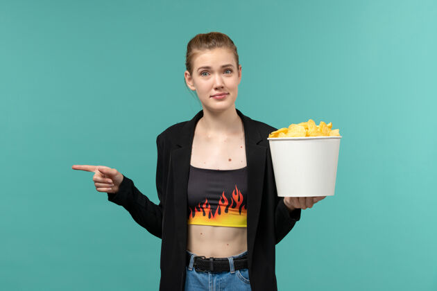 前面正面图：年轻女性手持薯片 在浅蓝色的表面上看电影土豆看衣服
