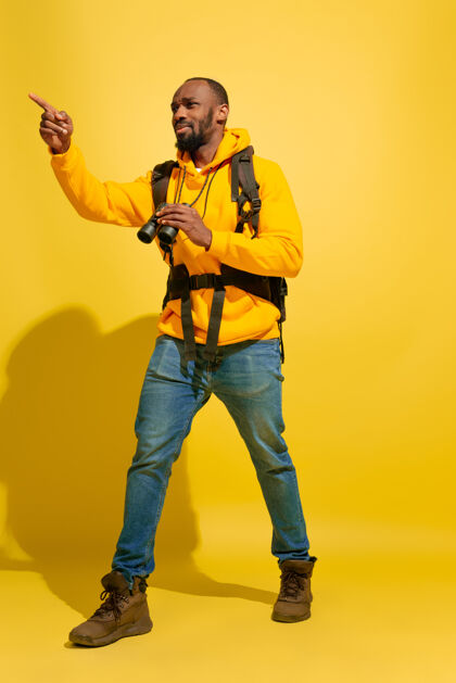 男性一个快乐的年轻游客的肖像 带着包和望远镜 隔离在黄色的工作室墙上旅行者举行度假村