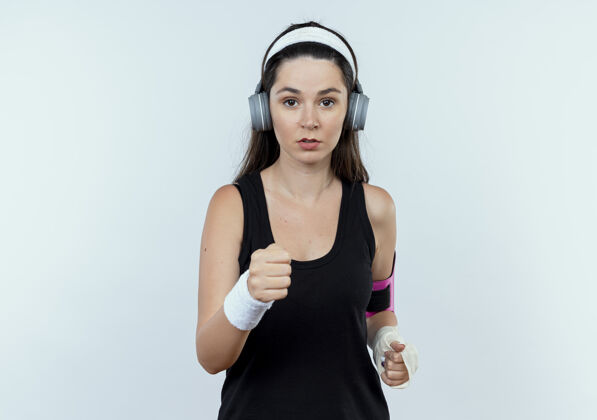 女士戴着头戴耳机和智能手机袖标的年轻健身女士站在白墙上严肃地锻炼身体严肃手机站立