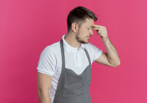 男人围裙上的理发师看起来很累 工作过度 站在粉红色的墙上闭着眼睛摸鼻子围裙摸着工作过度