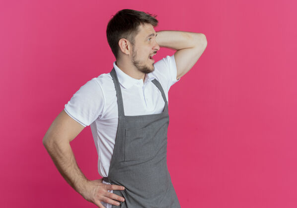 男人围着围裙的理发师站在粉红色的墙上 一边看一边困惑地抓着头站着理发师困惑
