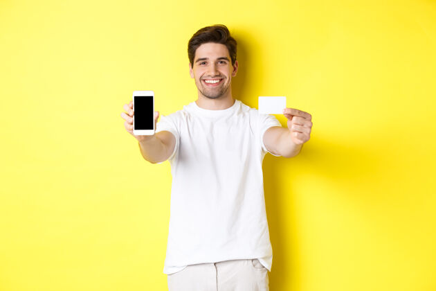 表情帅气的白人男模展示智能手机屏幕和信用卡 手机银行和网上购物概念 黄色背景模特移动男人