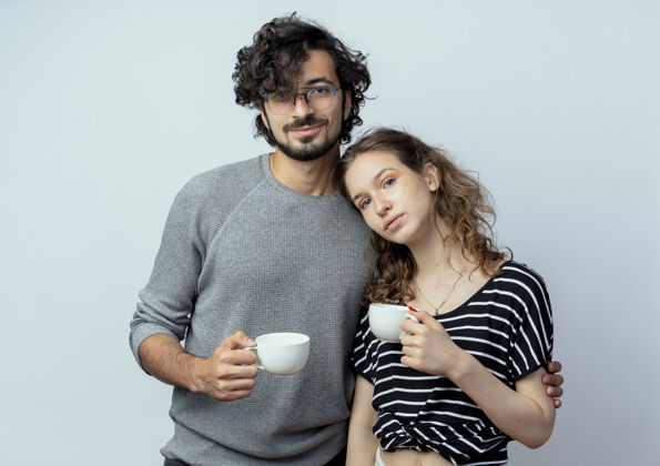积极年轻漂亮的情侣男女情投意合捧着咖啡杯在白墙上感受着积极的情感情绪情侣抱着