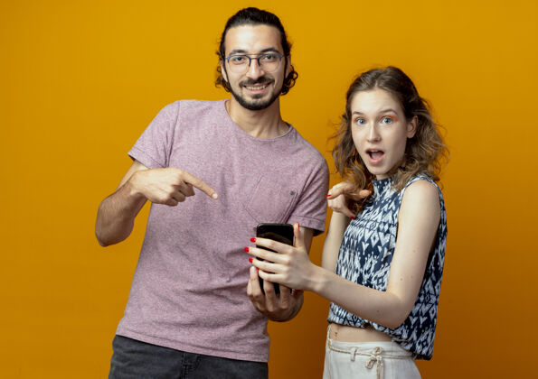 手指一对年轻夫妇拿着一部智能手机 用手指指着橙色的墙上 惊讶而高兴惊喜女人年轻