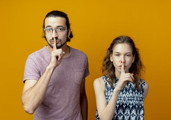 做这对年轻夫妇站在橘色的墙上 手指放在嘴唇上做着沉默的手势沉默手势站着