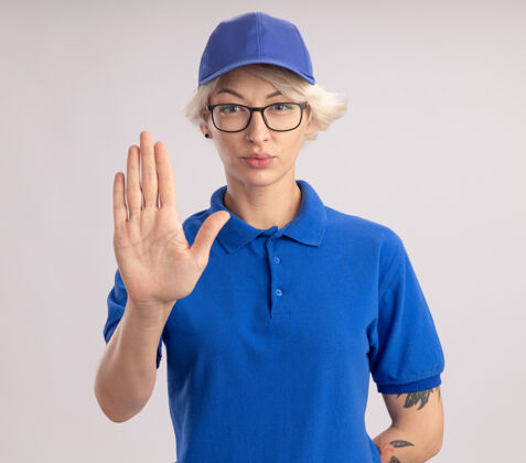 脸身穿蓝色制服 头戴鸭舌帽 表情严肃的年轻女送货员站在白色的墙上 摆出张开的手 做出停止的手势打开站立女人