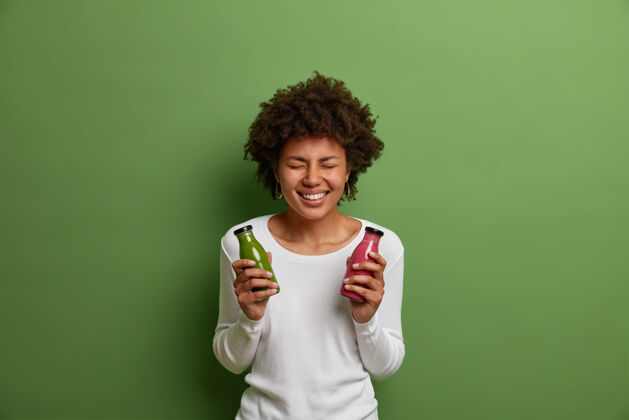 人室内拍摄的非常开心的微笑的女人拿着几瓶菠菜和草莓奶昔 喜欢喝排毒饮料 摆出一副新鲜饮料的姿势 隔离在绿色背景上欢呼生活方式非洲