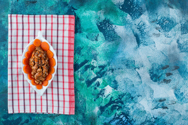 美味把切成片的胡萝卜和豆子放在茶巾上的盘子里 放在蓝色的背景上蔬菜可口烘焙