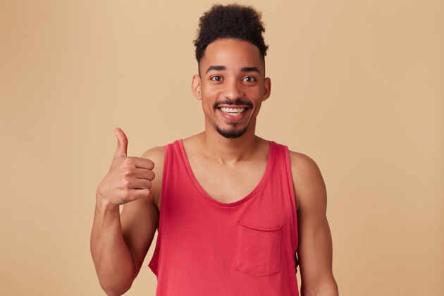独自英俊 快乐的非洲裔美国男性的肖像 非洲式发型和胡须穿着红色背心竖起大拇指的标志 都很好地覆盖了粉彩的米色墙壁外观胡须英俊