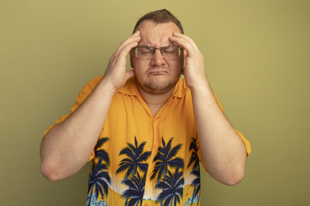 男人一个戴着眼镜 穿着橘色衬衫的男人站在绿色的墙上 看起来不舒服 摸着头 头痛得厉害头痛不舒服摸着