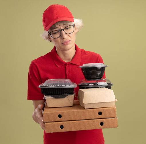 年轻年轻的送货员身穿红色制服 戴着帽子 戴着眼镜 手里拿着披萨盒和食品包 站在绿色的墙壁上 神情悲伤 疲惫 无聊地往下看盒子抱制服