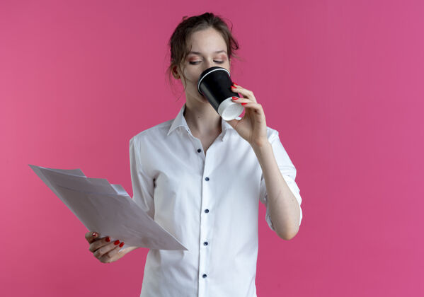 俄罗斯人年轻自信的金发俄罗斯女孩喝着咖啡看着纸页孤立在粉红色的空间与复印空间金发纸张饮料