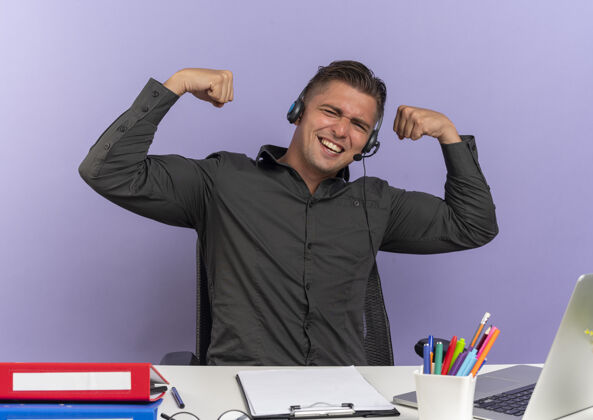 办公室年轻快乐的金发上班族戴着耳机坐在办公桌旁 拿着办公工具用笔记本电脑时态二头肌隔离在紫色空间和复印空间工人紧张工具