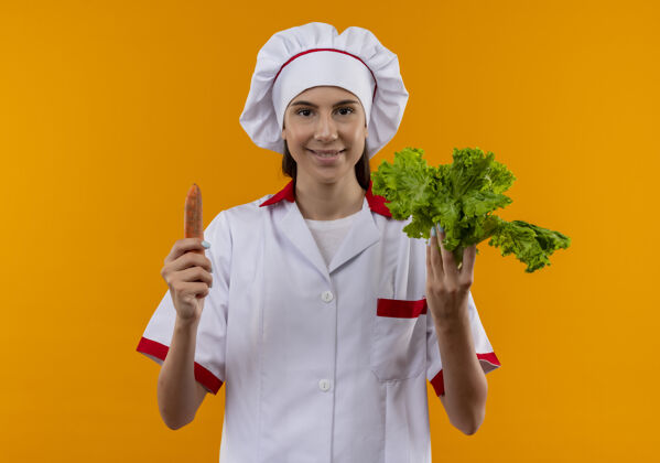 微笑身着厨师制服 面带微笑的白人年轻厨师女孩拿着胡萝卜和沙拉 隔离在橙色空间和复制空间年轻女孩烹饪