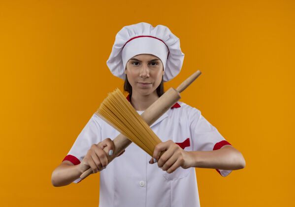 橙色年轻自信的白人厨师女孩穿着厨师制服 穿过擀面杖和一串意大利面 在橙色的空间和复制空间隔离卷复制信心