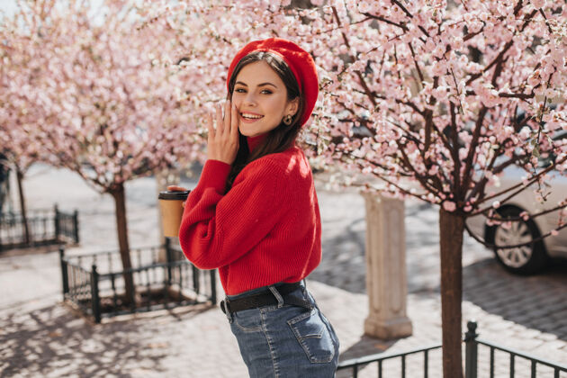 美女在樱花附近 一位乐观的女士穿着鲜艳的衣服 微笑着可爱一位穿着红毛衣 戴着红帽子的漂亮女士在城市花园里摆出一副好心情太阳镜帽子时尚