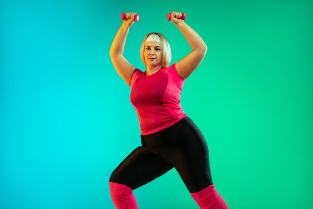 健身年轻的白人大号女模特在霓虹灯下的渐变绿色背景下训练用重量做锻炼运动的概念 健康的生活方式 积极的身体 平等加上运动专业