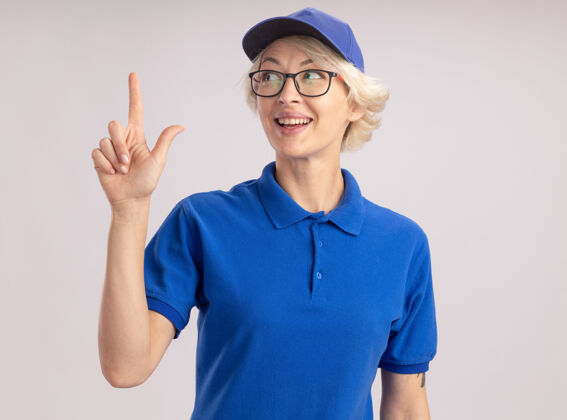 女人身穿蓝色制服 头戴鸭舌帽的年轻女送货员微笑着自信地指着站在白色墙壁上的食指手指帽子年轻