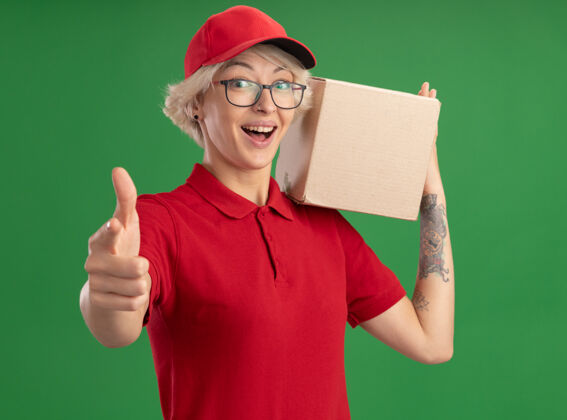 帽子年轻的送货员身穿红色制服 戴着帽子 戴着眼镜 手里拿着一个纸板箱 站在绿色的墙上 看起来很高兴 很兴奋 竖起大拇指年轻展示拇指