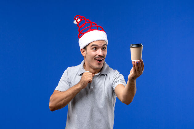 咖啡正面图年轻男子拿着咖啡杯站在蓝色墙上新年男子节日感慨圣诞节情绪前面