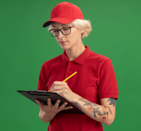 铅笔身穿红色制服 戴着帽子的年轻送货员戴着一副眼镜 写字板上写着空白页 铅笔上写着一张严肃的脸站在绿色的墙上剪贴板帽子穿