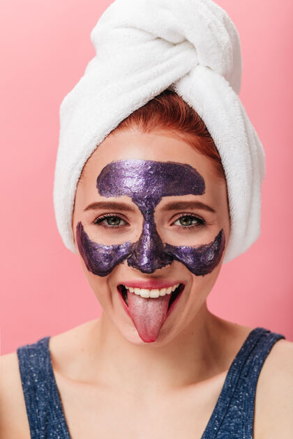 健康有趣的女孩在做水疗时露出舌头的前视图带着面具在粉色背景上摆姿势的极乐女人的摄影棚照片治疗完美的皮肤护理