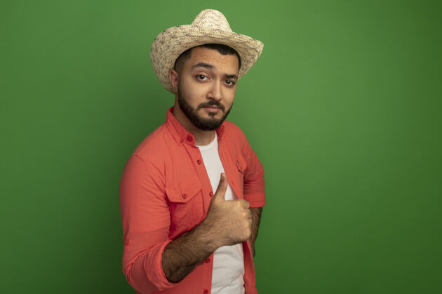 拇指年轻的留着胡子的男人穿着橙色衬衫 戴着夏天的帽子 站在绿色的墙上竖起大拇指年轻人帽子男人