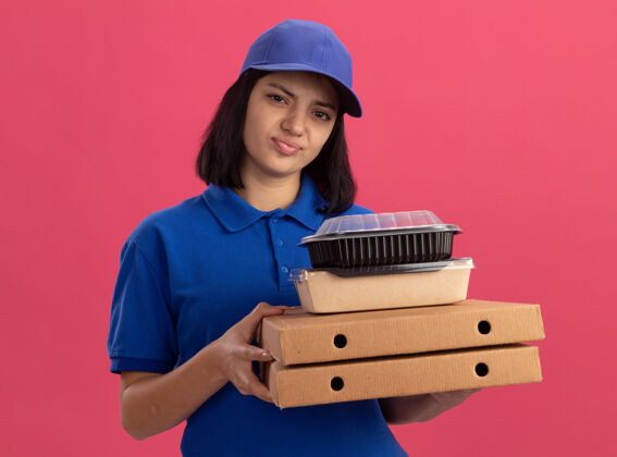 盒子站在粉红色的墙上 一个穿着蓝色制服 戴着帽子 拿着比萨饼盒和食品包 看起来很不高兴的悲伤的年轻送货女孩年轻包装站着