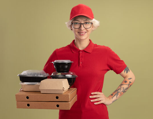 自信年轻的送货员身穿红色制服 戴着帽子 戴着眼镜 手里拿着披萨盒和食品包 自信地微笑着 手臂放在臀部 站在绿色的墙上年轻人胳膊女人