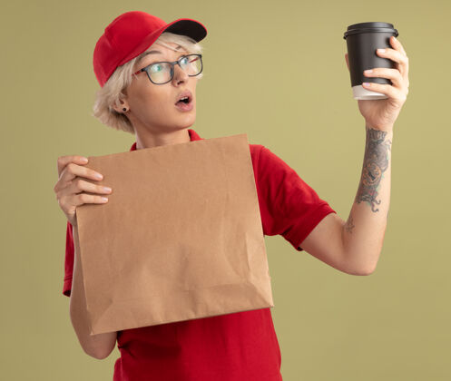 穿年轻的送货员身穿红色制服 戴着帽子 戴着眼镜 手里拿着纸包 看着手中的咖啡杯 站在绿色的墙上 惊讶又惊讶手拿着包装