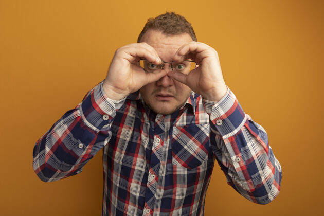 站一个戴着眼镜和格子衬衫的男人站在橙色的墙上做着双眼手势望远镜眼镜手势