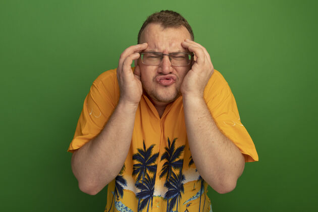 男人一个戴着眼镜 穿着夏威夷衬衫 手放在头上 站在绿色的墙上 看上去既困惑又担心的男人眼镜站着手