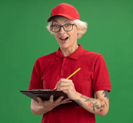 送货年轻的送货员身穿红色制服 戴着一顶帽子 戴着一副眼镜 写字板上有空白的纸和铅笔 站在绿色的墙上 微笑着 高兴着 惊讶地写着什么穿空白眼镜
