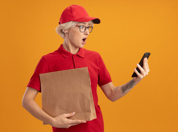 拿着年轻的送货员身穿红色制服 戴着帽子 戴着眼镜 手里拿着纸包 站在橙色的墙上 惊讶地看着自己的智能手机惊喜穿上眼镜