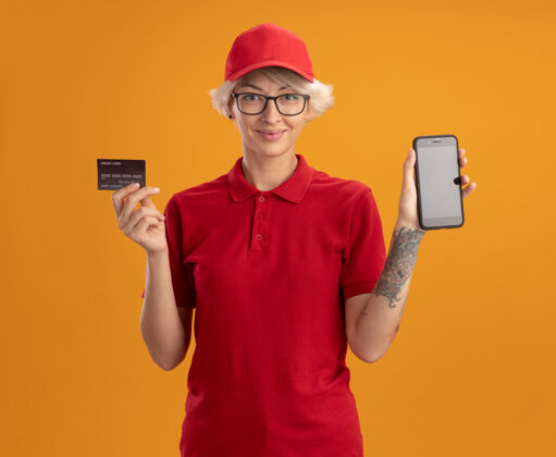 显示身着红色制服 戴着眼镜的年轻女送货员站在橙色的墙上 脸上露出智能手机和信用卡的微笑帽子送货手机