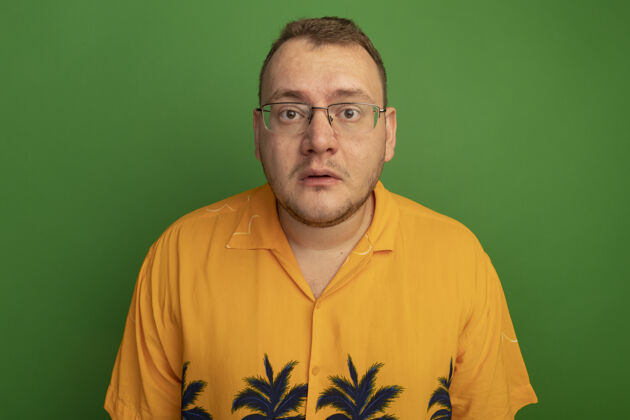 非常一个戴着眼镜 穿着夏威夷衬衫的男人站在绿色的墙上 感到困惑和焦虑站着焦虑男人
