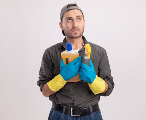 年轻年轻的保洁员穿着休闲服 戴着橡胶手套 手里拿着喷雾瓶和清洁用品 站在白色的墙上困惑地抬头望去男人橡胶喷雾
