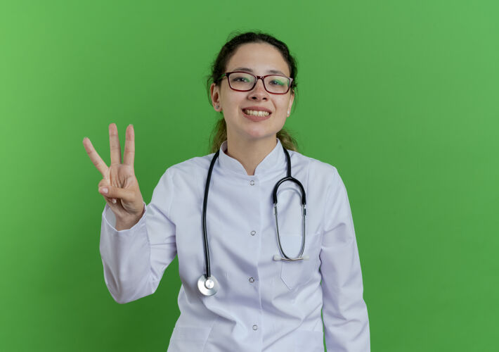 微笑面带微笑的年轻女医生 穿着医用长袍 戴着听诊器和眼镜 三只手被隔离在绿墙上 留有复印空间医生年轻听诊器