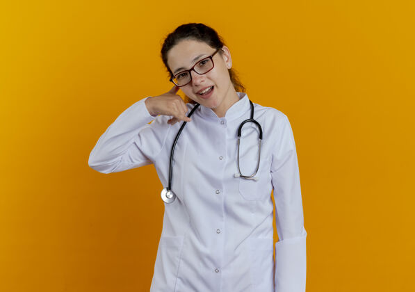 眼镜微笑着的年轻女医生 穿着医用长袍 戴着听诊器 戴着眼镜 在橙色的墙上显示着电话的手势听诊器女性医生