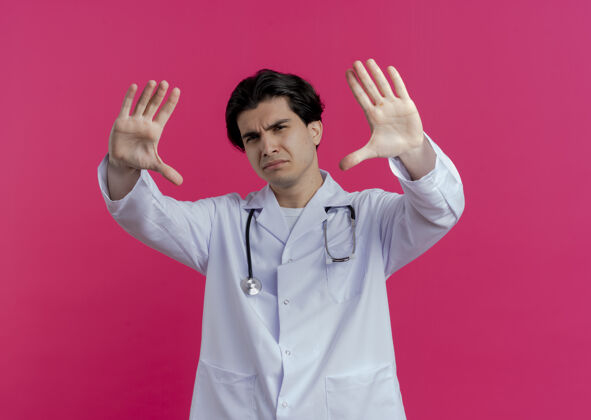 男性穿着医用长袍和听诊器的年轻男性医生在粉红色的墙上做停止手势 并留有复印空间不愉快长袍手势