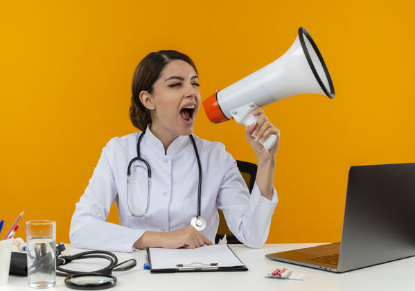 医生年轻的女医生穿着医用长袍和听诊器坐在办公桌旁 拿着医疗工具和笔记本电脑 在黄色的墙上大声喊叫喊叫工具办公桌